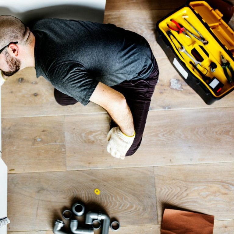 Person kneeling on a wooden floor, surrounded by tools and plumbing materials.