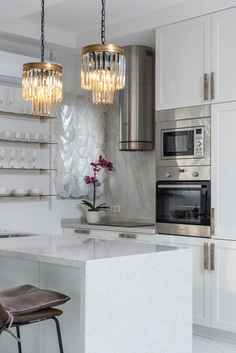 A modern kitchen with a waterfall countertop on a white cabinet island, modern appliances, and crystal light fixtures.