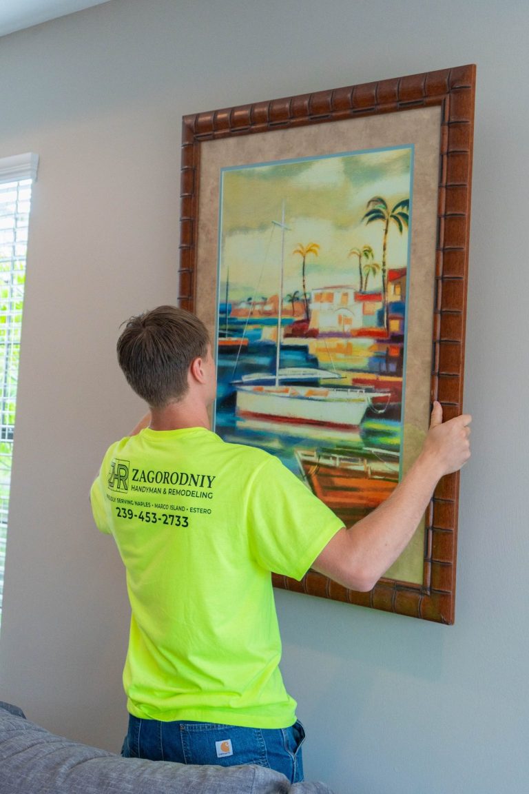 A technician in a neon shirt hangs a framed coastal painting on a light-colored wall in Naples.