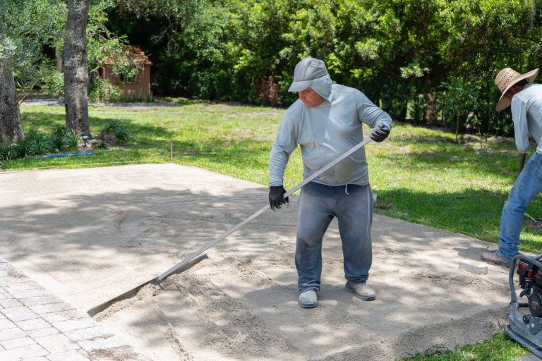 A worker using a long tool to level a pad of sand in a grey shirt and hat in North Naples.