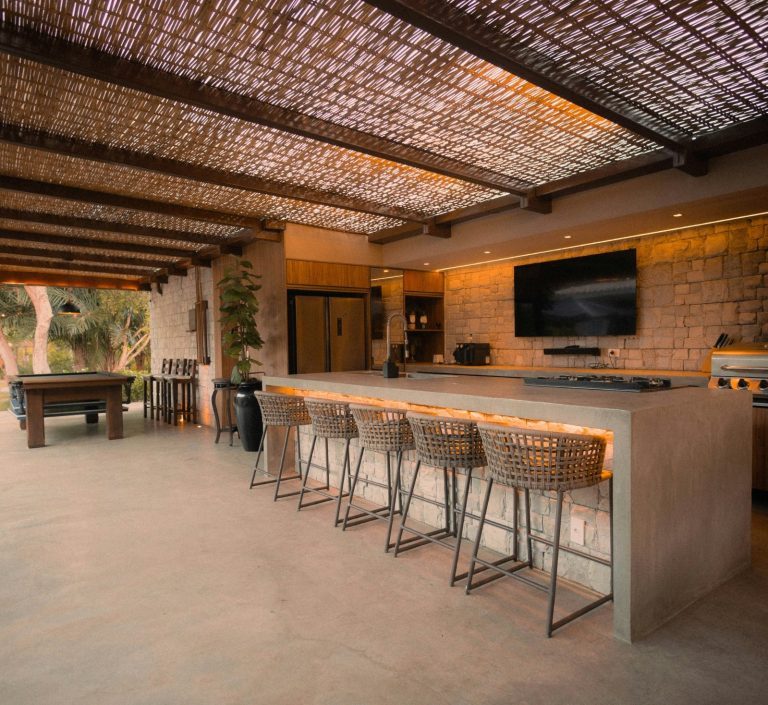 Outdoor kitchen and bar area with high stools, a countertop, and a television, under a shaded roof in Marco Island, FL.